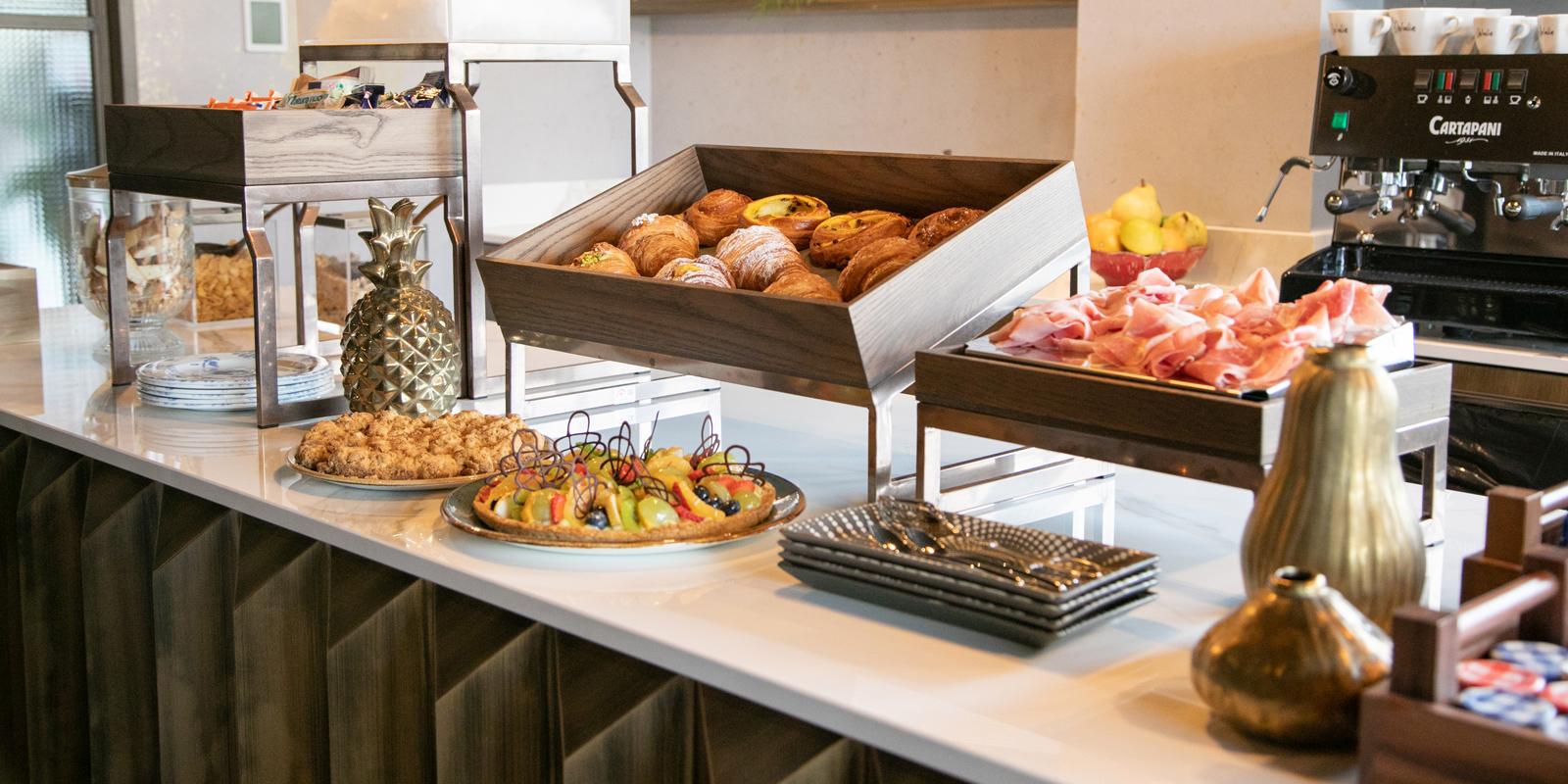 Breakfast buffet with pastries, fruit tart, ham, and coffee machine in a modern setting.