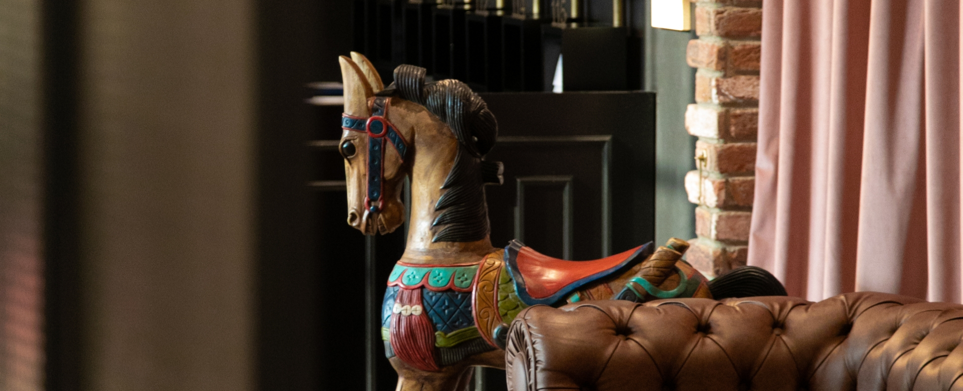 Wooden carousel horse beside a leather couch and pink curtains.