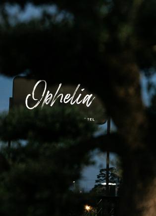 Sign for 'Ophelia' hotel, partially obscured by trees, seen during twilight.