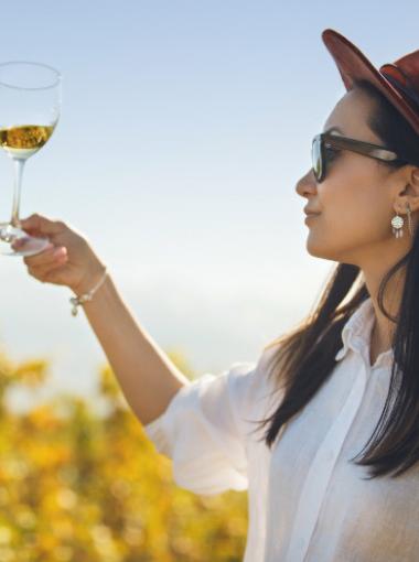 Woman in hat and sunglasses holding a wine glass in a vineyard.