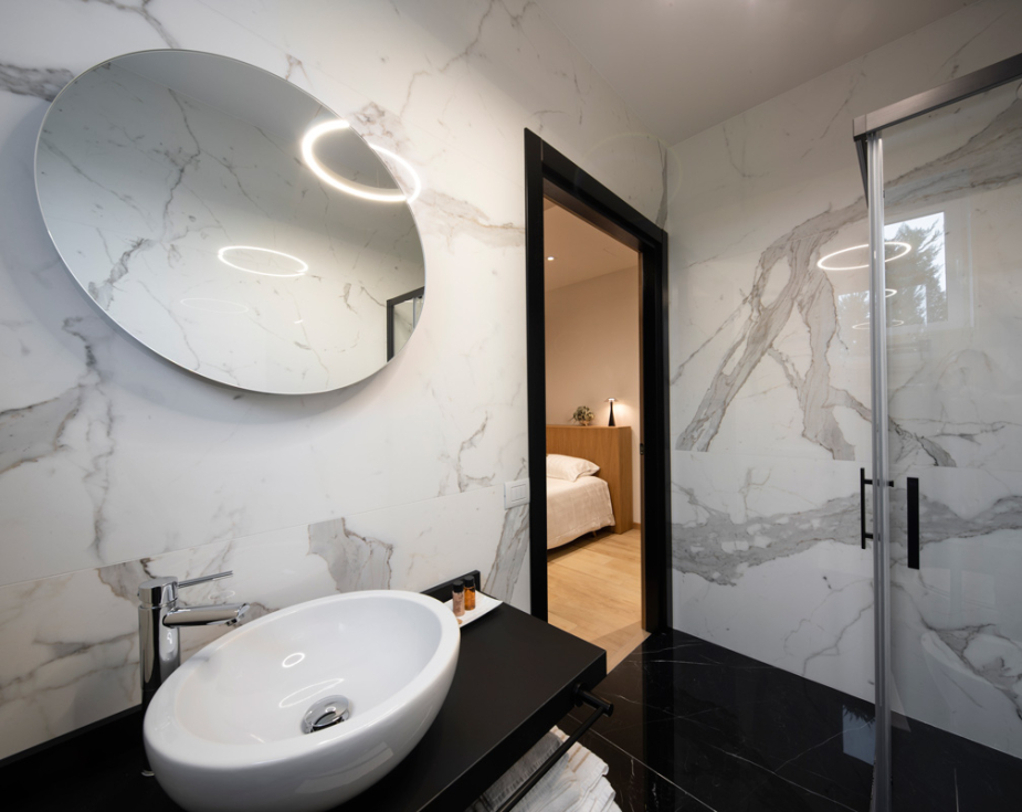 Modern bathroom with marble walls, circular mirror, and a view into the bedroom.