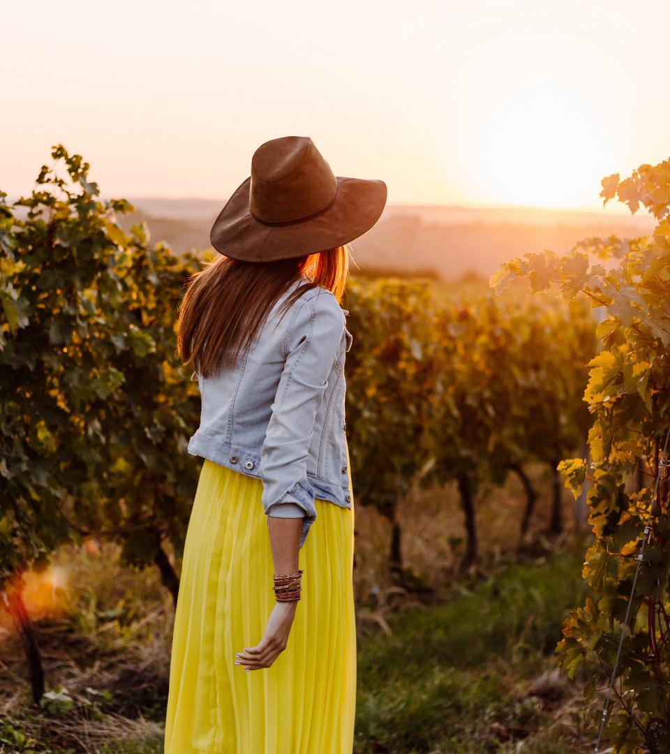 Donna con cappello in un vigneto al tramonto, indossa giacca di jeans e gonna gialla.