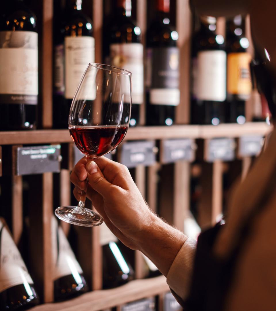 A person holding a glass of red wine in front of wine bottles.